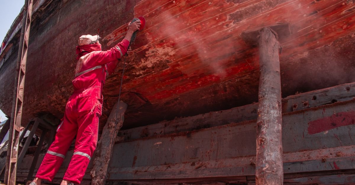 Worker in overalls stripping the hull of a ship with abrasive equipment in a shipyard