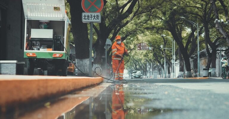 Municipal worker using a power washer to clean a wet city pavement, spray reflecting off the soaked surface