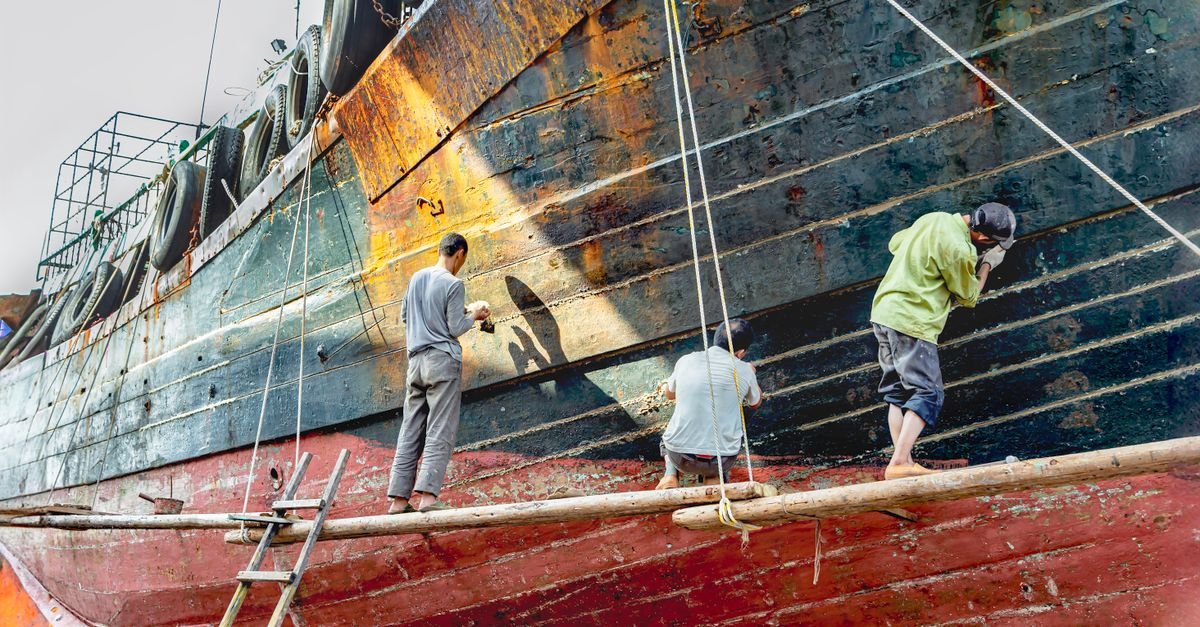 Three workers refurbishing a ship's hull in a yard, standing at the hull side with the bottom exposed