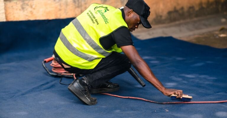 A worker in a safety vest scrubbing a blue outdoor carpet with a stiff brush before rinsing