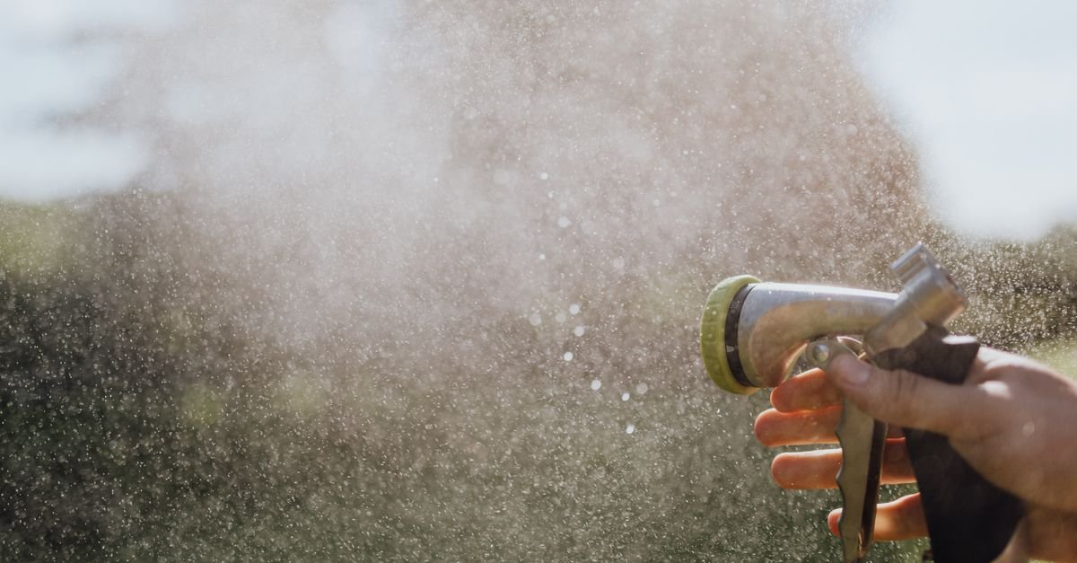 A gloved hand holding a garden hose nozzle producing a soft fan of water outdoors