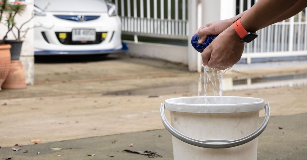 Man cleaning car with sponge and bucket in driveway, showcasing everyday car maintenance routine
