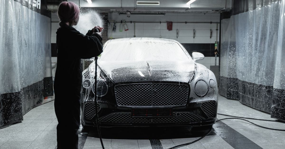 A black Bentley being washed with soap at an indoor car wash facility by a woman in a black sweater