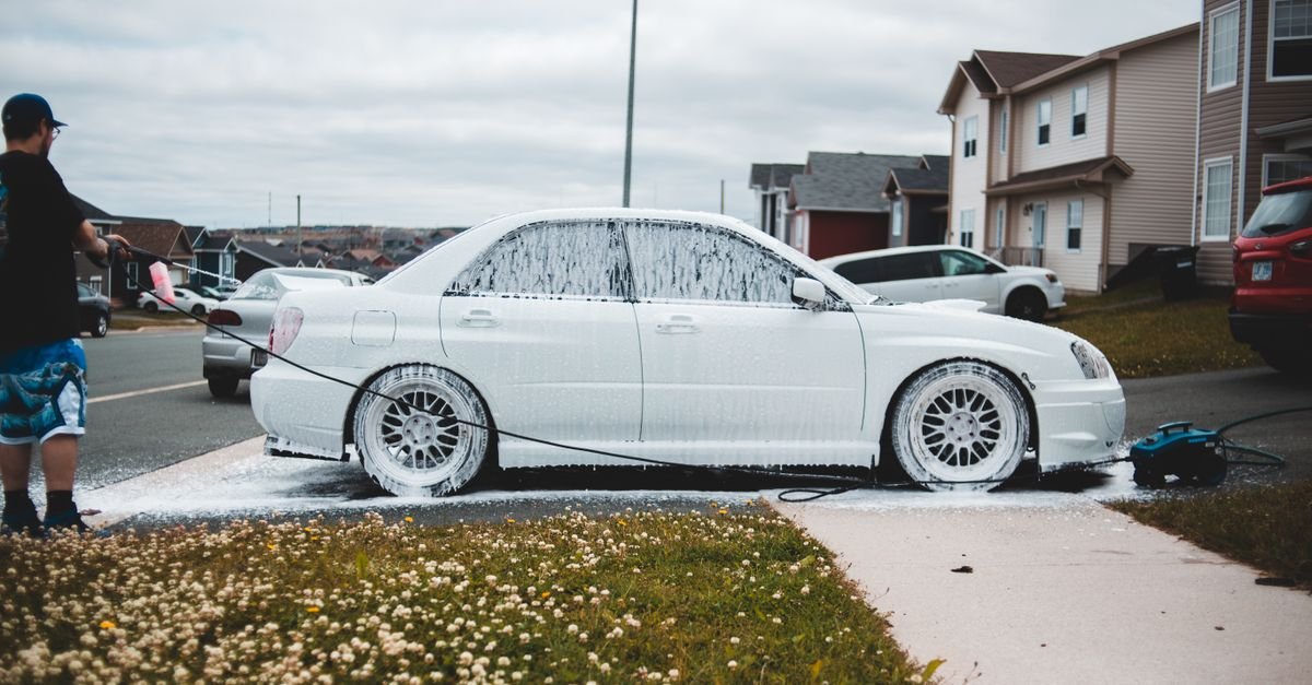 Foamed car on a driveway being washed by a man with a pressure washer during the day