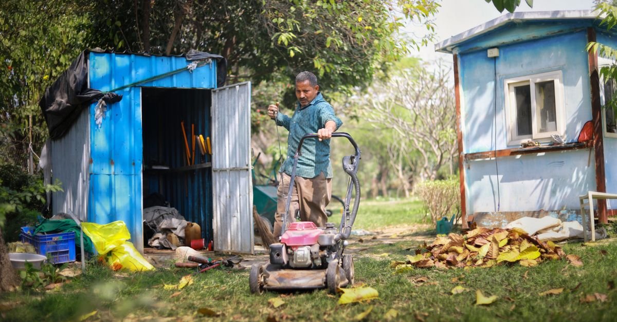 Gardener mowing a lawn beside a blue garden shed with leaf piles in a lush garden