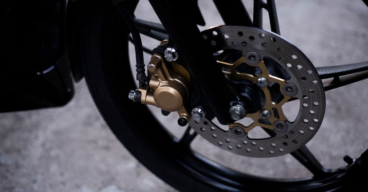 Close-up of a motorcycle front brake disc and caliper, the drilled rotor and pads fully exposed on the outside of the wheel