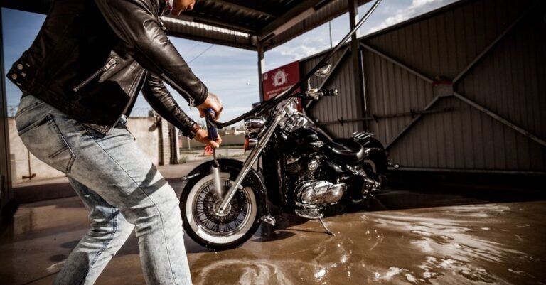 Man rinsing a motorcycle with a garden hose at an outdoor wash bay on a sunny day