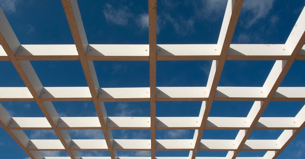 Low-angle view from beneath a timber pergola, the underside of the rafters and slats against open sky