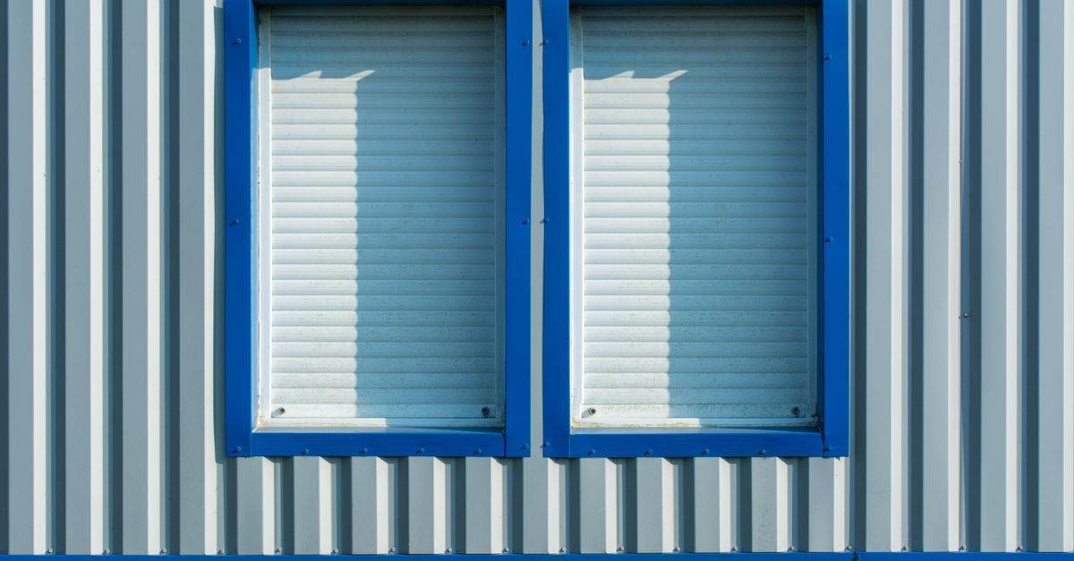 Close-up of a corrugated metal clad wall with blue-framed windows and shutters set into it