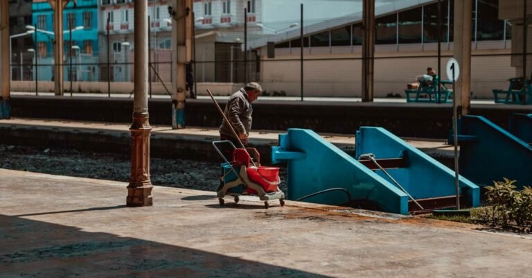 A worker scrubbing a long concrete platform with cleaning equipment under a station canopy