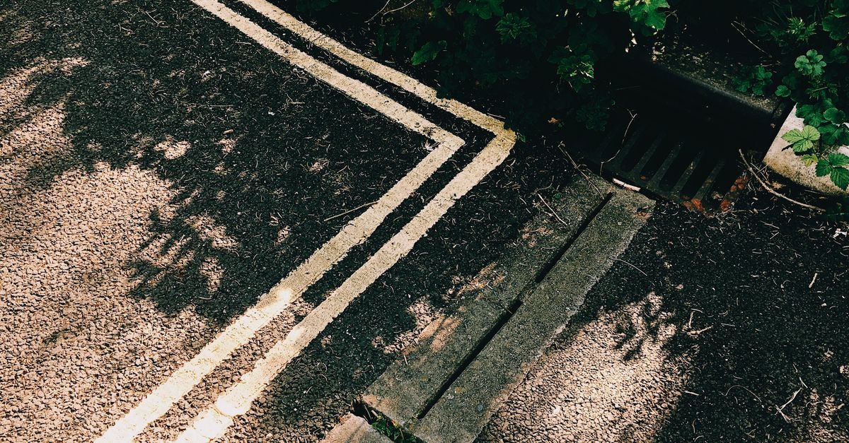 Sunlit asphalt road with white line markings beside a metal sewer grate at the kerb