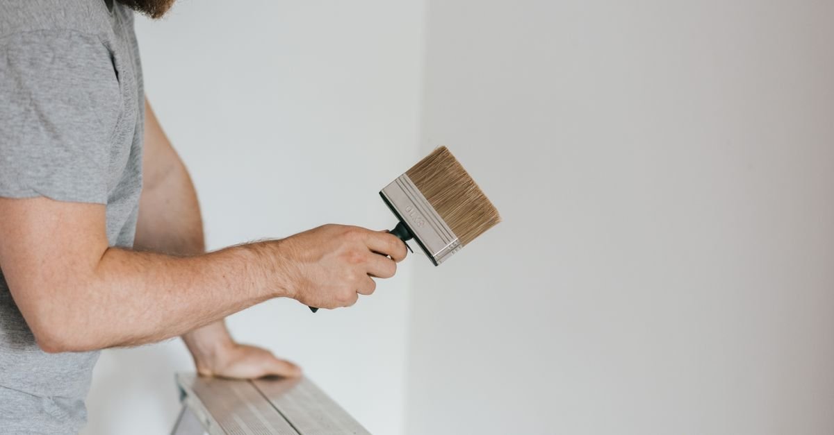A worker on a ladder holding a brush against an exterior wall, working the surface by hand