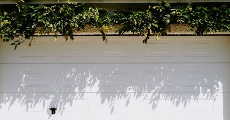 A white raised-panel steel sectional garage door in full sun, leaf shadows falling across the horizontal section joints