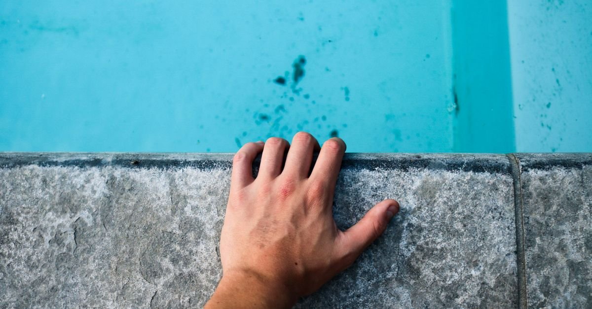 Close-up of a hand resting on the concrete edge of a swimming pool with clear blue water below