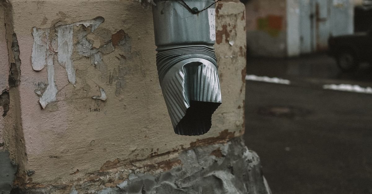 Weathered metal downpipe on a wall with flaking paint, the kind of fitting roof runoff drains through