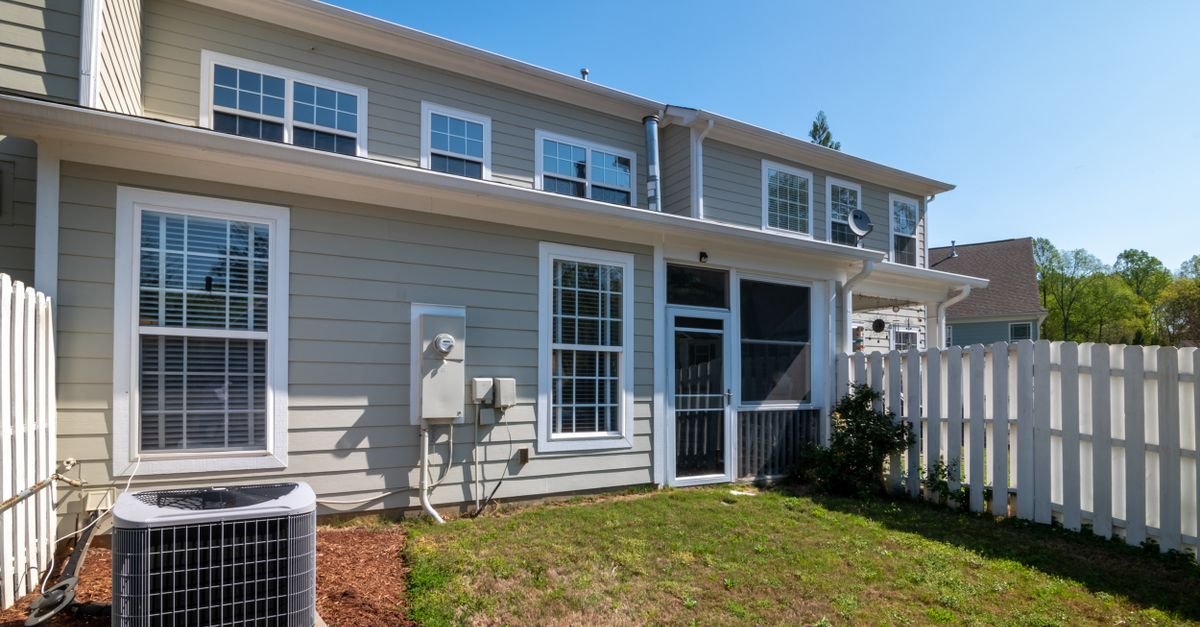 A grey house with white trim and an air conditioning condenser standing on the ground against the wall