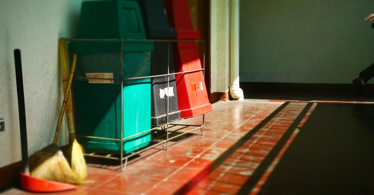 Wheeled bins standing beside cleaning tools in a bright corridor, brush handles leaning against the wall