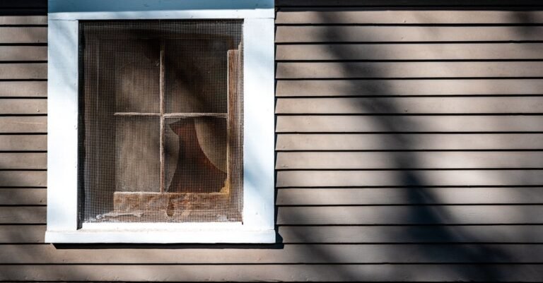 Exterior of a rustic wooden house with an insect screen over the window casting shadows in low sunlight