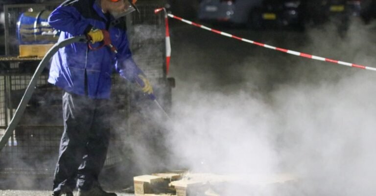 Worker in protective gear operating a commercial pressure washer amid steam on a night job site