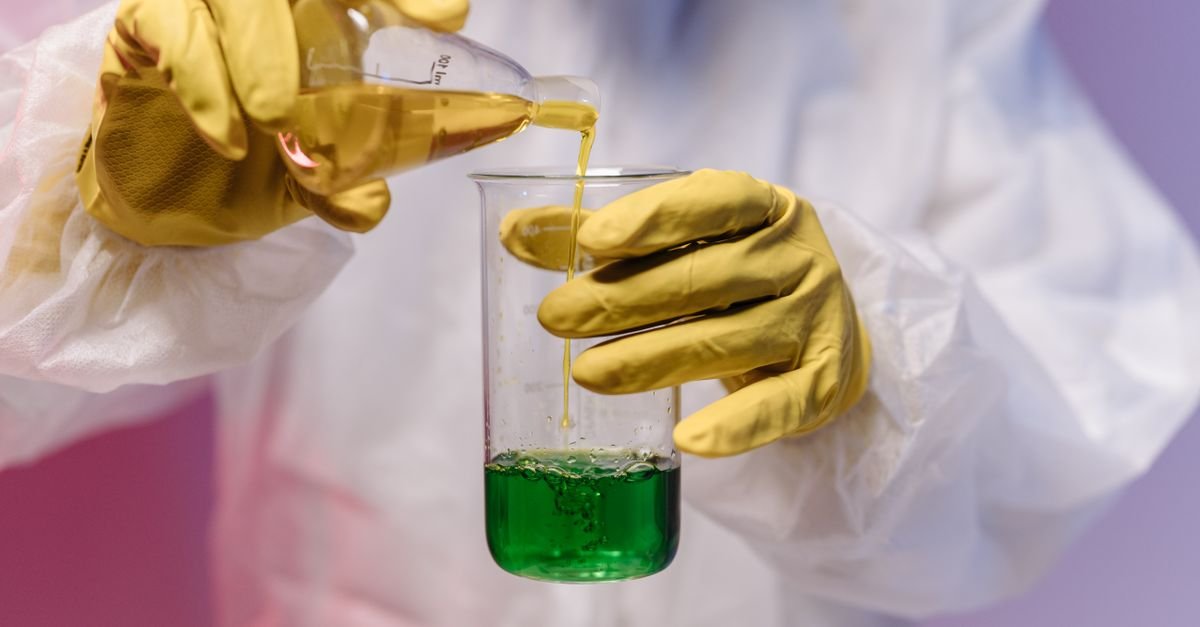 Gloved hands in protective clothing carefully pouring a chemical solution into a measuring vessel