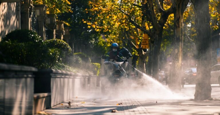 A municipal worker spraying water across a concrete sidewalk on a sunny day