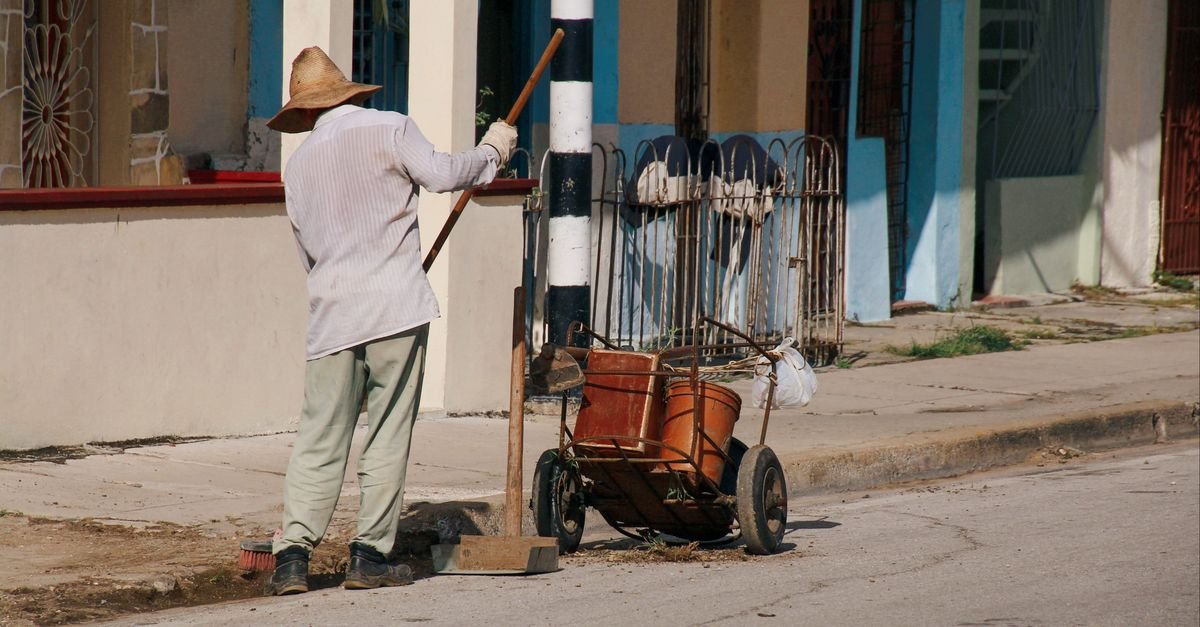 A worker sweeping a city pavement by hand with a broom and a handcart