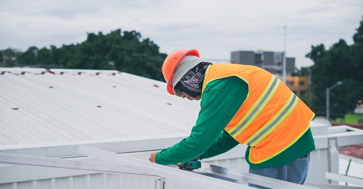 A worker in a hard hat and fall-protection harness kneeling on a residential rooftop