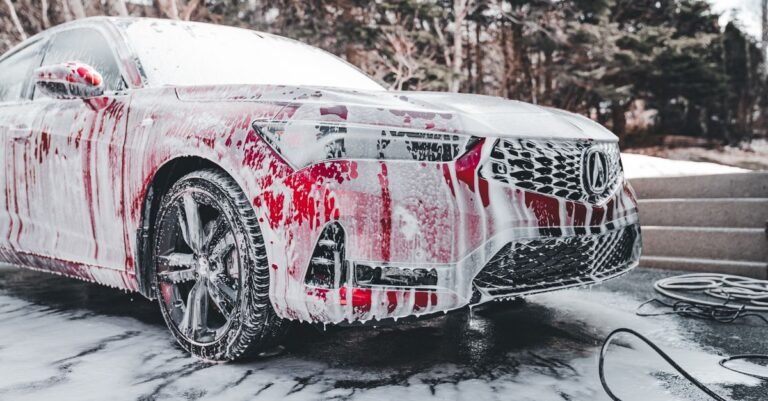 Red car completely covered in thick foam cannon suds on an outdoor driveway