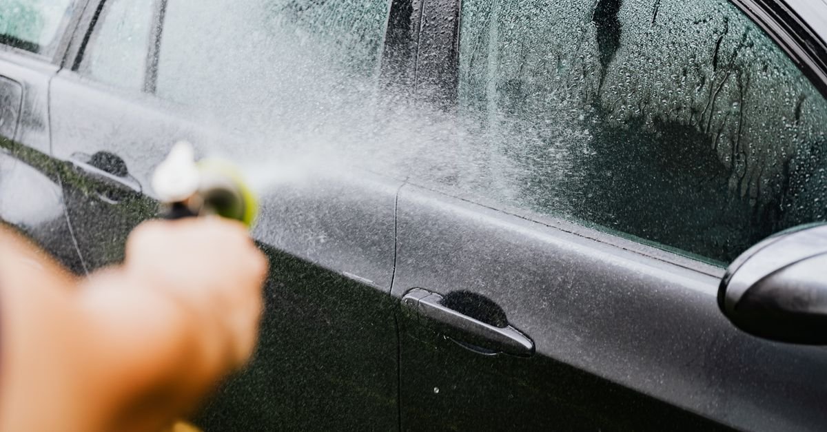 Car being rinsed with an ordinary garden hose outdoors, water spraying across the paintwork
