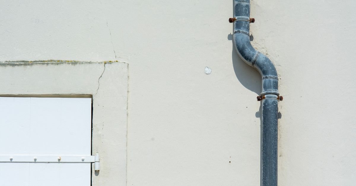 A grey metal downspout running down the outside wall of a house, showing the offset at the base