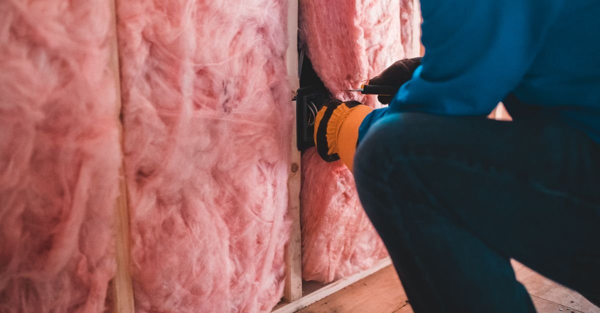 A gloved worker pressing pink mineral wool insulation into place between roof joists