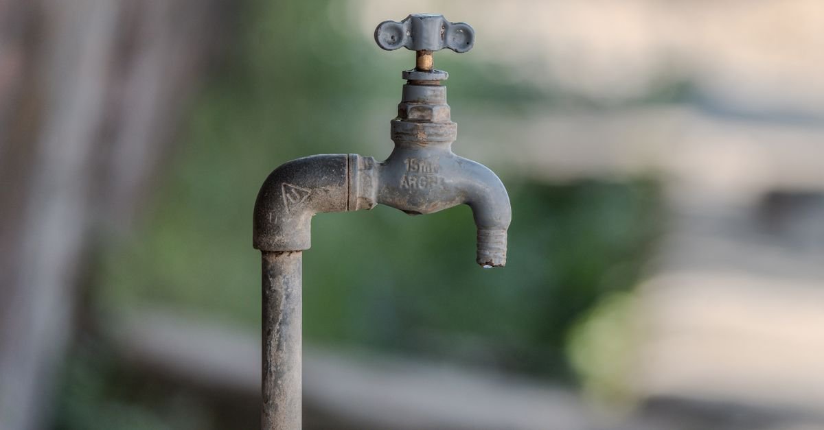 A weathered outdoor water faucet on a wall with a green garden blurred behind it