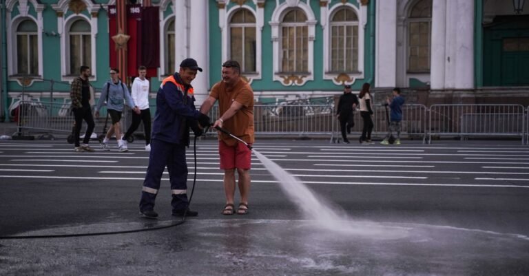 Two workers in high-visibility clothing washing down a city street with a hose in front of a historic stone building