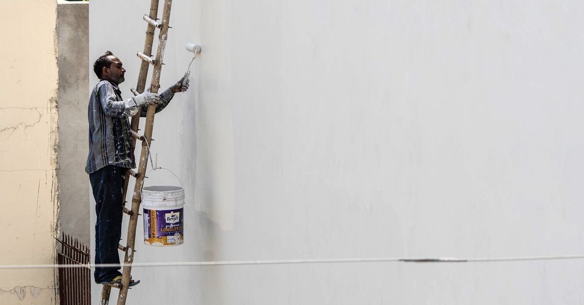 A man on a ladder reaching up to work a roller across a tall exterior wall outdoors