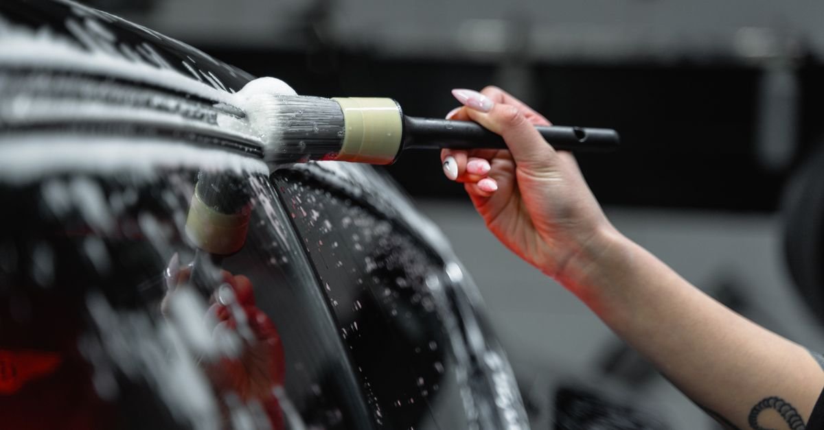 A hand working a soapy wash brush across a wet painted panel, foam lifting the dirt film off the surface