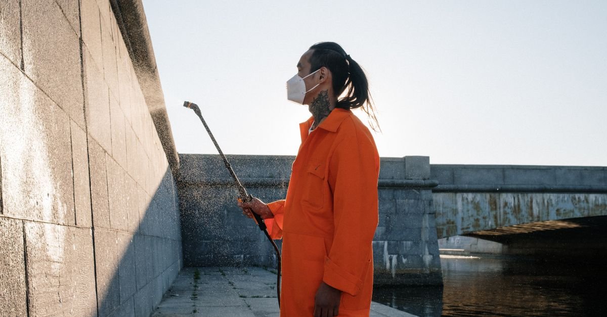 Worker in protective gear pressure washing a concrete wall with a fan-tip wand