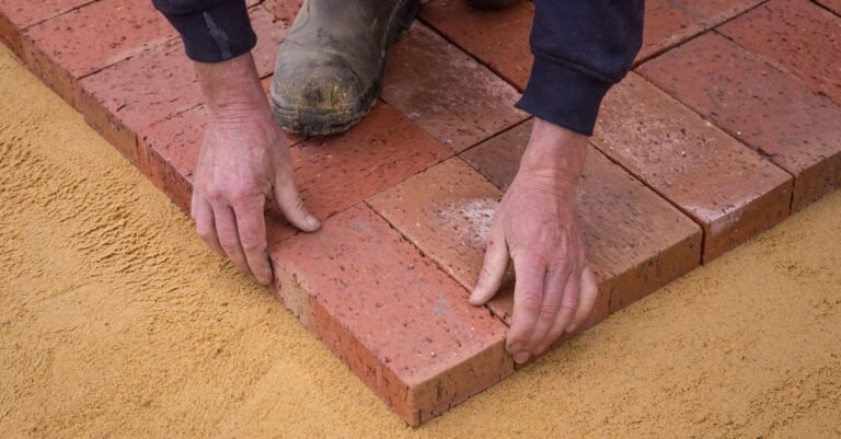 Worker's hands setting clay brick pavers onto a sand bed