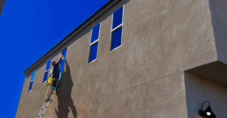 Man on a ladder cleaning the exterior windows of a building under a clear blue sky