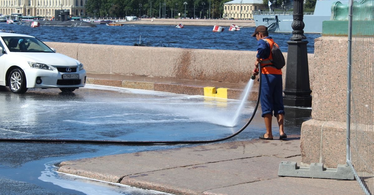 A city worker rinsing a wet paved surface with a hose, water running away downhill