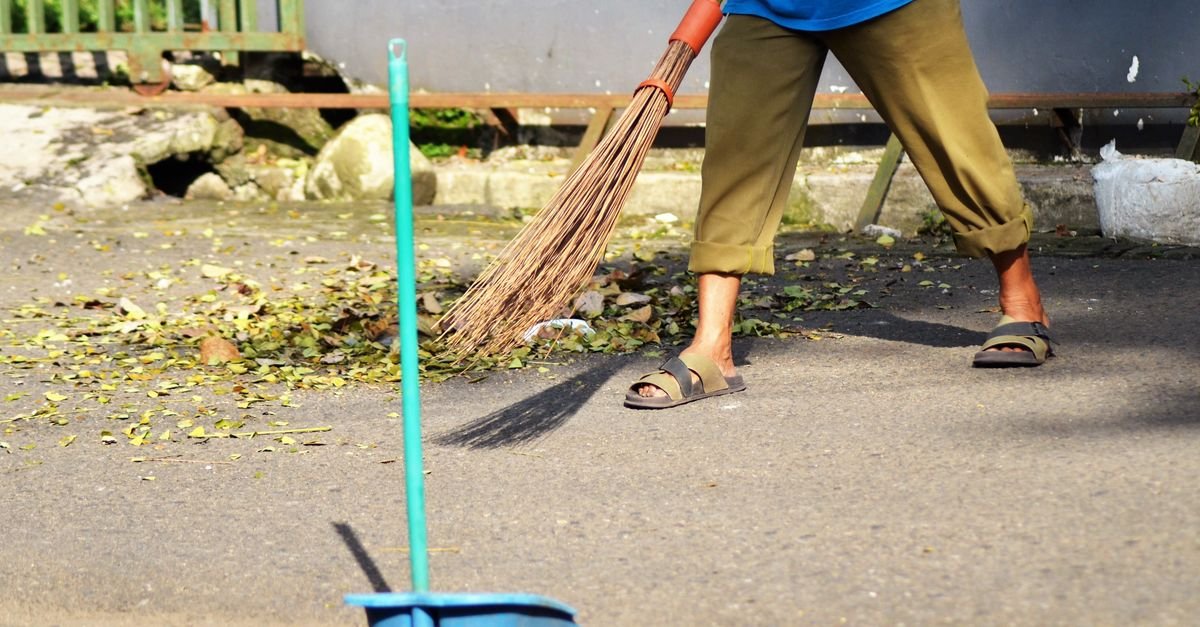 A person sweeping fallen leaves off a paved surface with a stiff broom