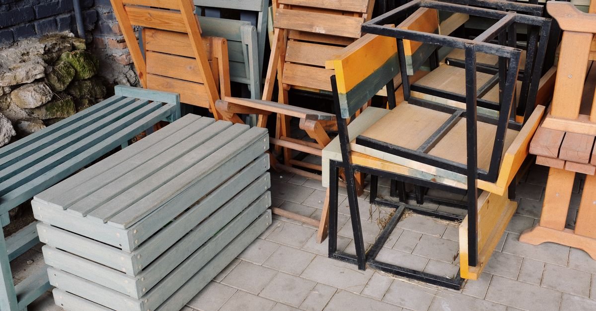 Wooden benches, chairs and metal frames stacked together outside on a paved area