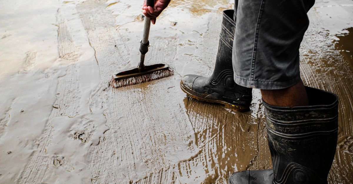 Worker in rubber boots scrubbing a wet concrete surface with a stiff push broom