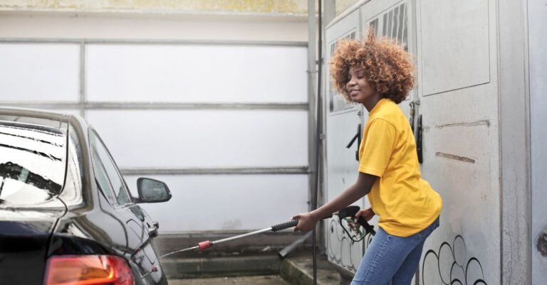 Woman in a yellow shirt using a jet washer to rinse a black car on a driveway