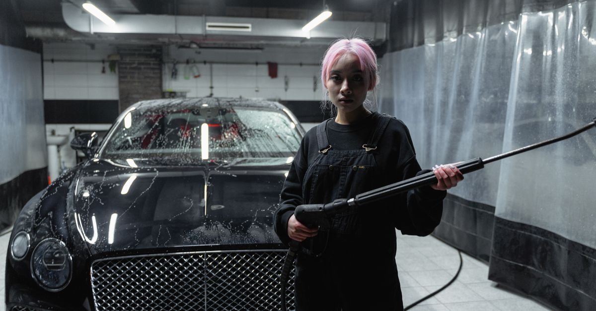 A person cleaning a dark car with a pressure washer in a garage, the everyday duty cycle that wears an unloader out