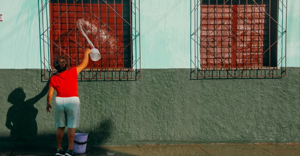 A woman in a red shirt washing the exterior of a green house by hand with a bucket and water