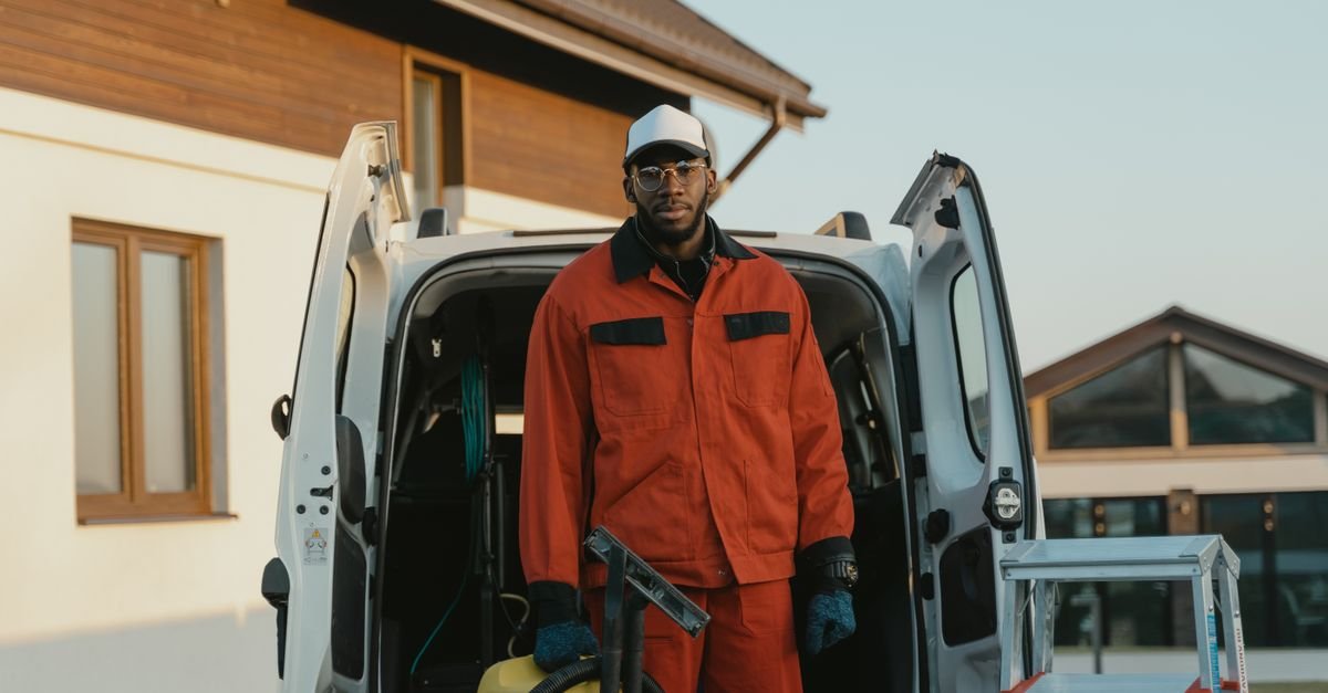 A worker in uniform with cleaning equipment beside a van outside a building before starting a job