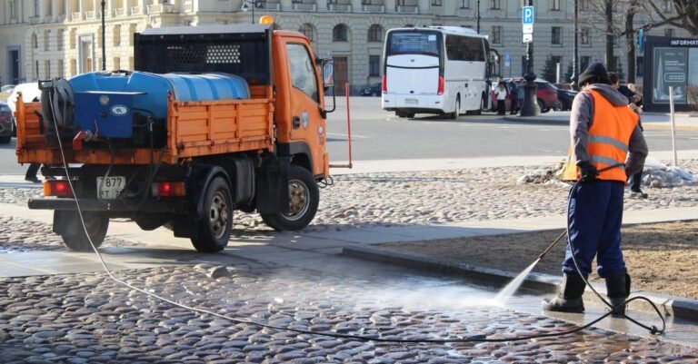 A city worker directing a high-pressure jet across a paved street surface in sunlight