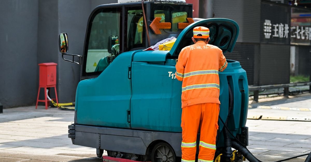 A street cleaner in uniform pushing an enclosed cleaning machine along an urban sidewalk