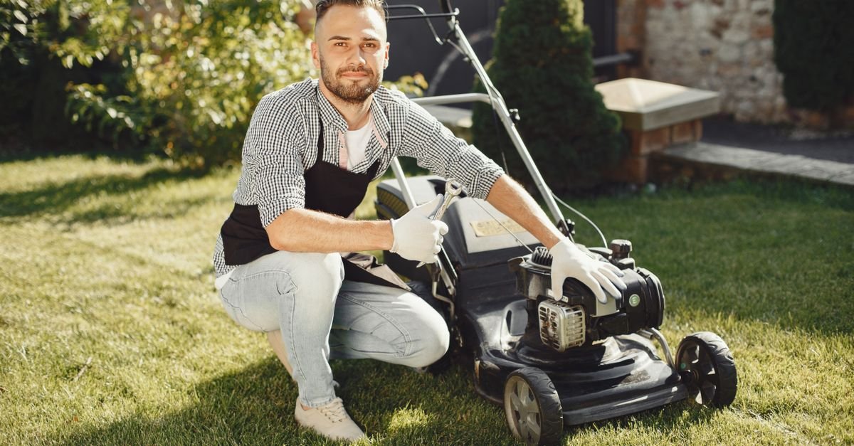 A man in gloves servicing a petrol lawn mower on a sunny lawn
