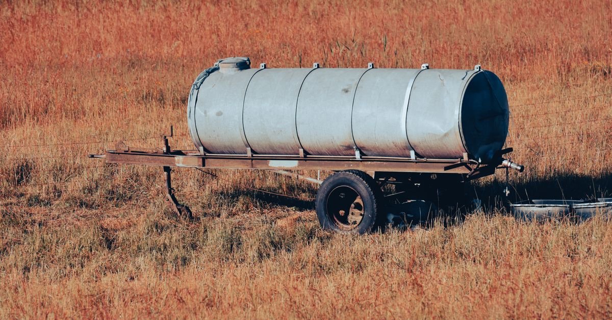 A metal water storage tank on wheels standing in a dry grassy field, the kind of gravity-fed supply that sits below the pump inlet